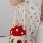 A toddler with red hair in two buns stands beside a white cylindrical pedestal, wearing a white sleeveless romper with small blue floral prints and holding a mushroom-shaped shoulder bag.