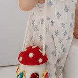 A toddler with red hair in two buns stands beside a white cylindrical pedestal, wearing a white sleeveless romper with small blue floral prints and holding a mushroom-shaped shoulder bag.