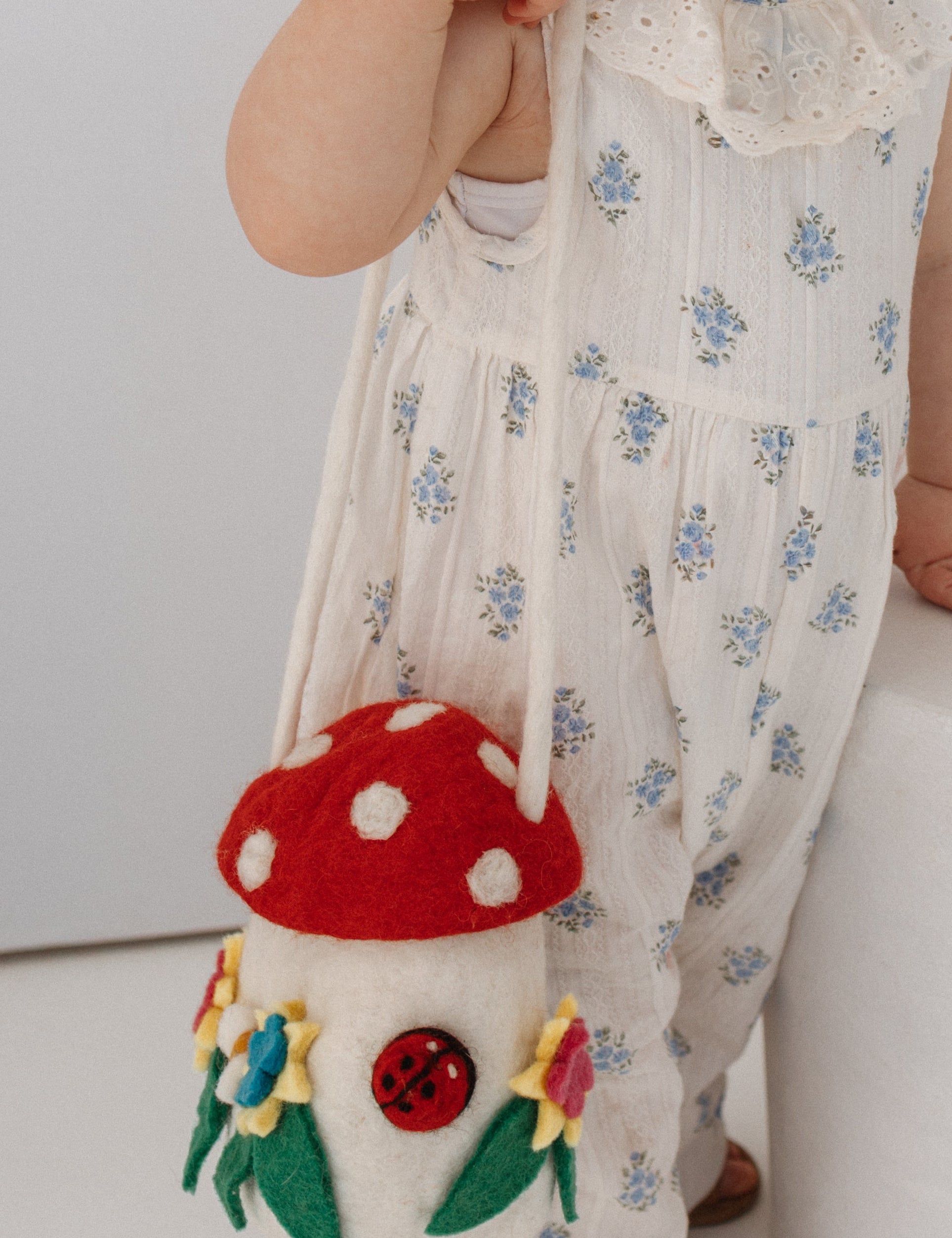 A toddler with red hair in two buns stands beside a white cylindrical pedestal, wearing a white sleeveless romper with small blue floral prints and holding a mushroom-shaped shoulder bag.