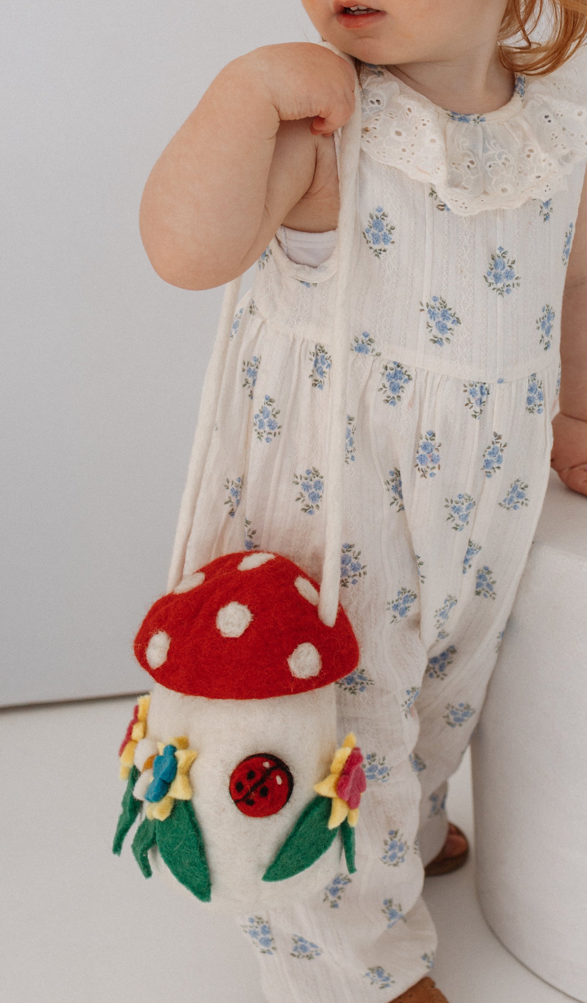 A toddler with red hair in two buns stands beside a white cylindrical pedestal, wearing a white sleeveless romper with small blue floral prints and holding a mushroom-shaped shoulder bag.