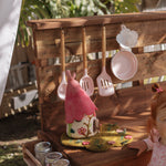 A young child in a pink dress stands beside a wooden outdoor play kitchen set on grass, with hanging utensils, small accessories, and a garden fence in the background. Fairy play, mud kitchen , fairy garden