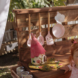 A young child in a pink dress stands beside a wooden outdoor play kitchen set on grass, with hanging utensils, small accessories, and a garden fence in the background. Fairy play, mud kitchen , fairy garden