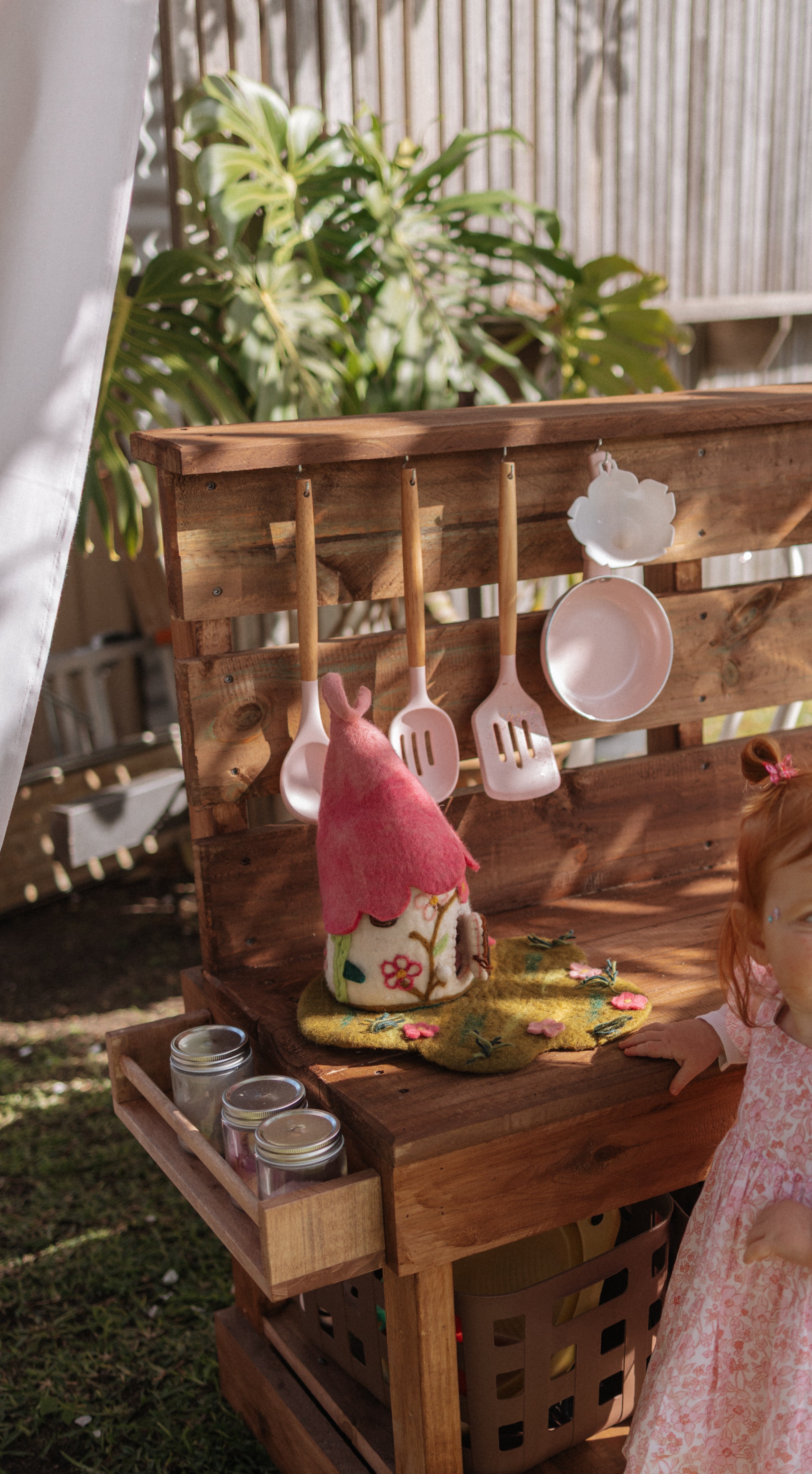A young child in a pink dress stands beside a wooden outdoor play kitchen set on grass, with hanging utensils, small accessories, and a garden fence in the background. Fairy play, mud kitchen , fairy garden