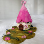A handmade felt fairy house with a bright pink pointed roof, a cream-colored body decorated with floral patterns, and a green felt ground base with small pink flowers.