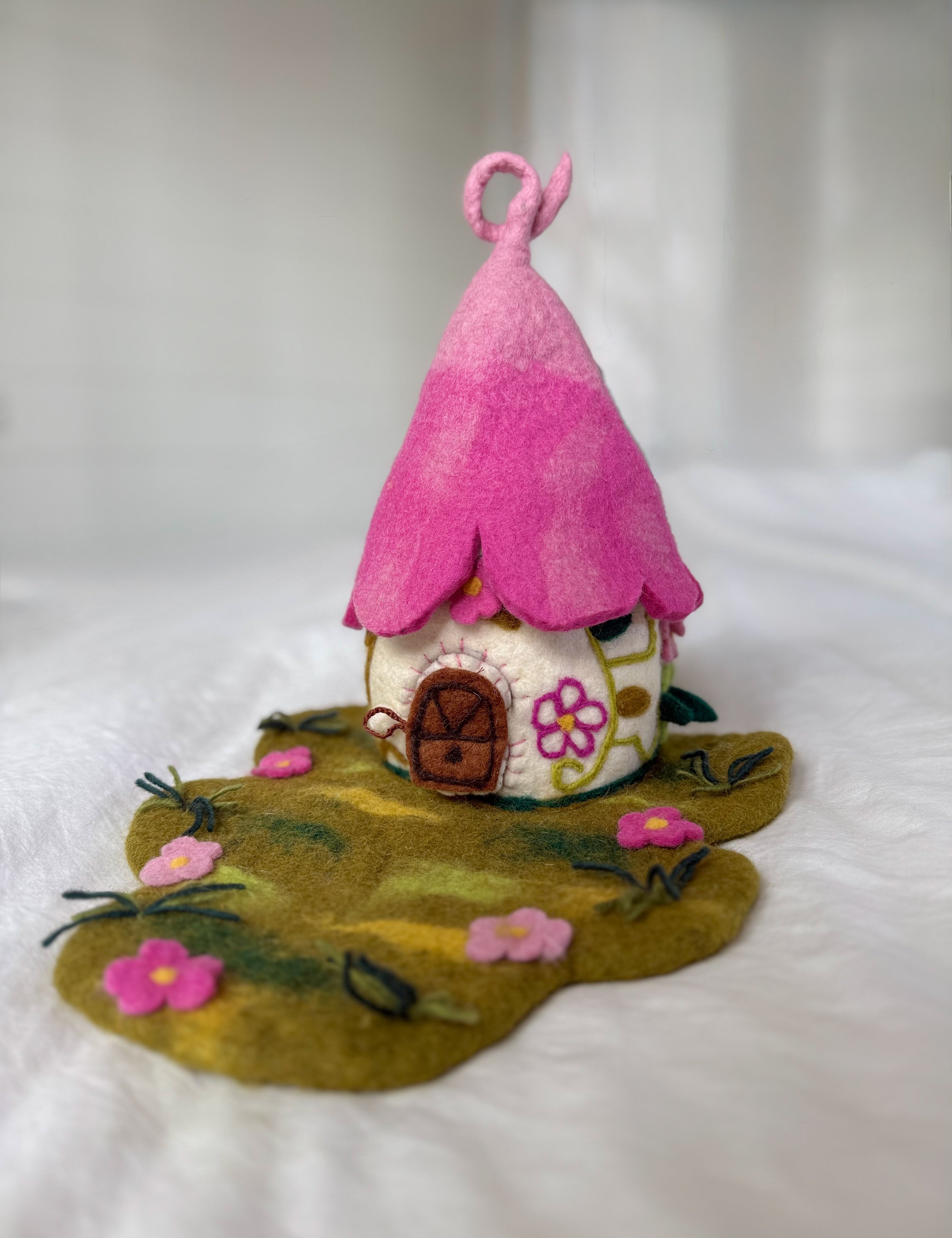 A handmade felt fairy house with a bright pink pointed roof, a cream-colored body decorated with floral patterns, and a green felt ground base with small pink flowers.