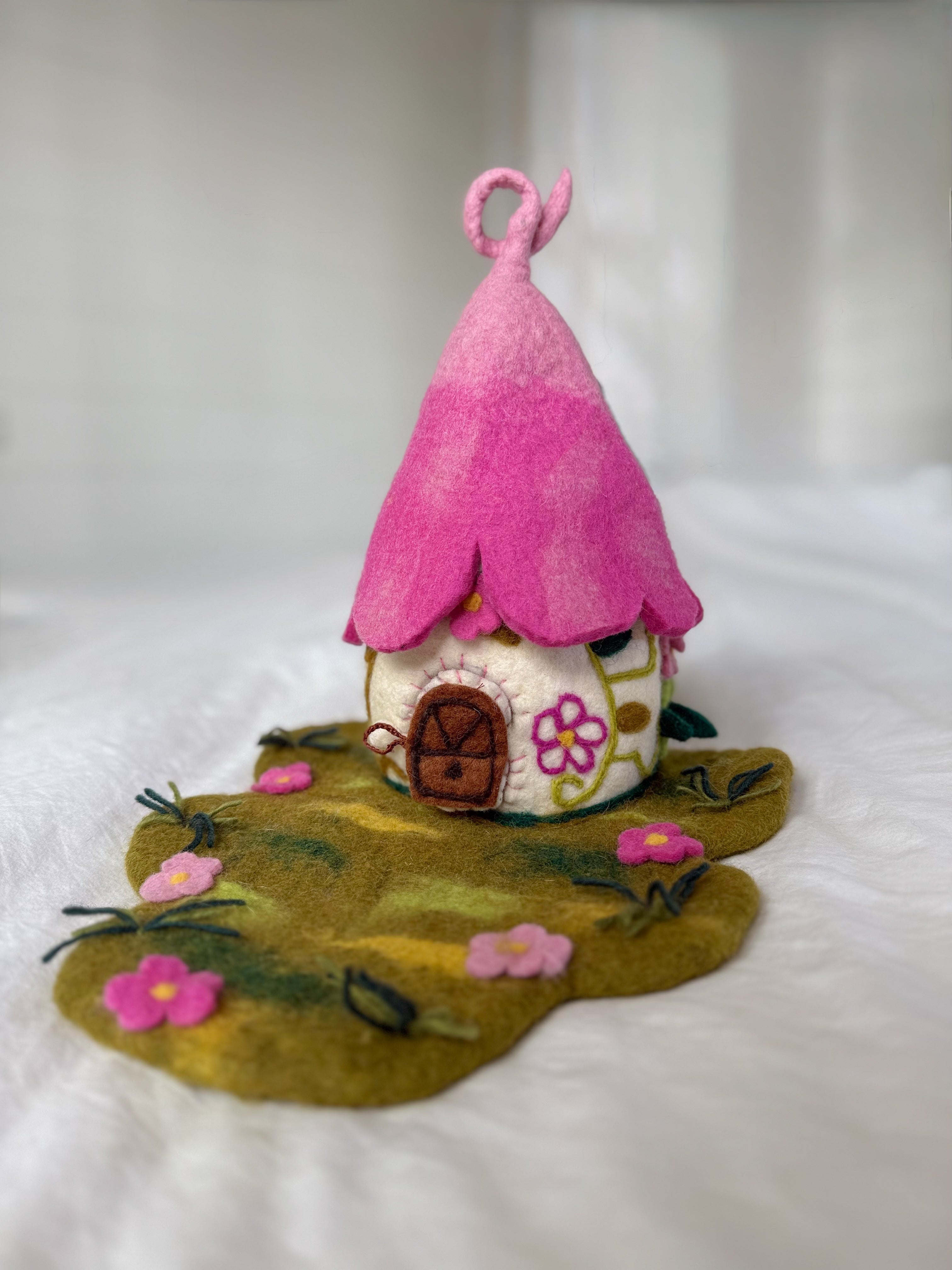 A handmade felt fairy house with a bright pink pointed roof, a cream-colored body decorated with floral patterns, and a green felt ground base with small pink flowers.