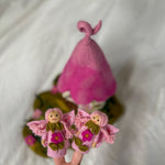 A pink felt fairy-themed product set featuring two small felt fairy figures with wings and flowers in the foreground, and a larger pink felt hat or hat-like structure behind them on a soft white fabric background.