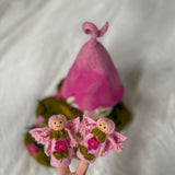 A pink felt fairy-themed product set featuring two small felt fairy figures with wings and flowers in the foreground, and a larger pink felt hat or hat-like structure behind them on a soft white fabric background.