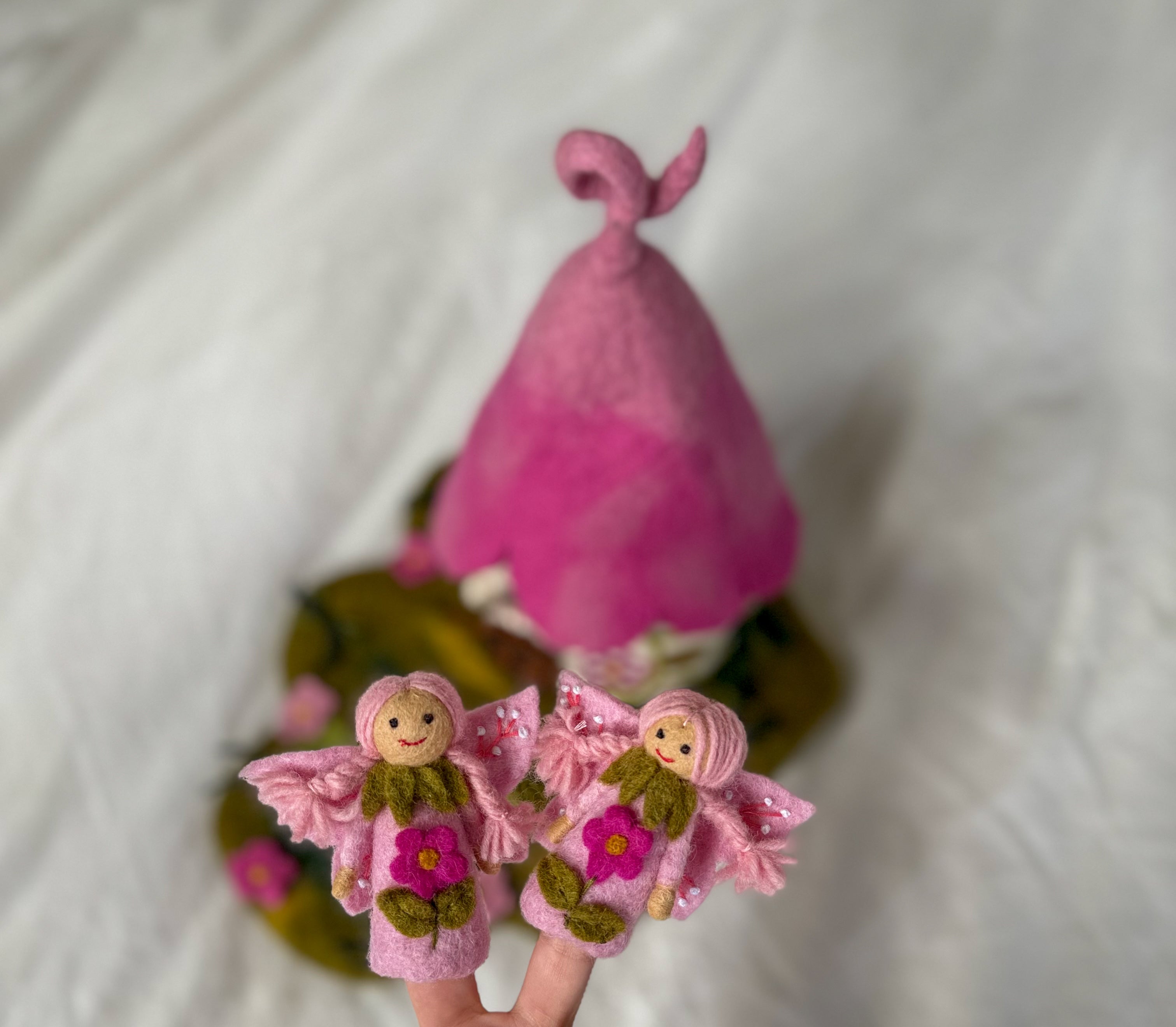A pink felt fairy-themed product set featuring two small felt fairy figures with wings and flowers in the foreground, and a larger pink felt hat or hat-like structure behind them on a soft white fabric background.
