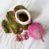 A felted wool baby bootie in the shape of a flower, shown with its matching pink felted wool hood and small pink floral accessories on a white fabric background.