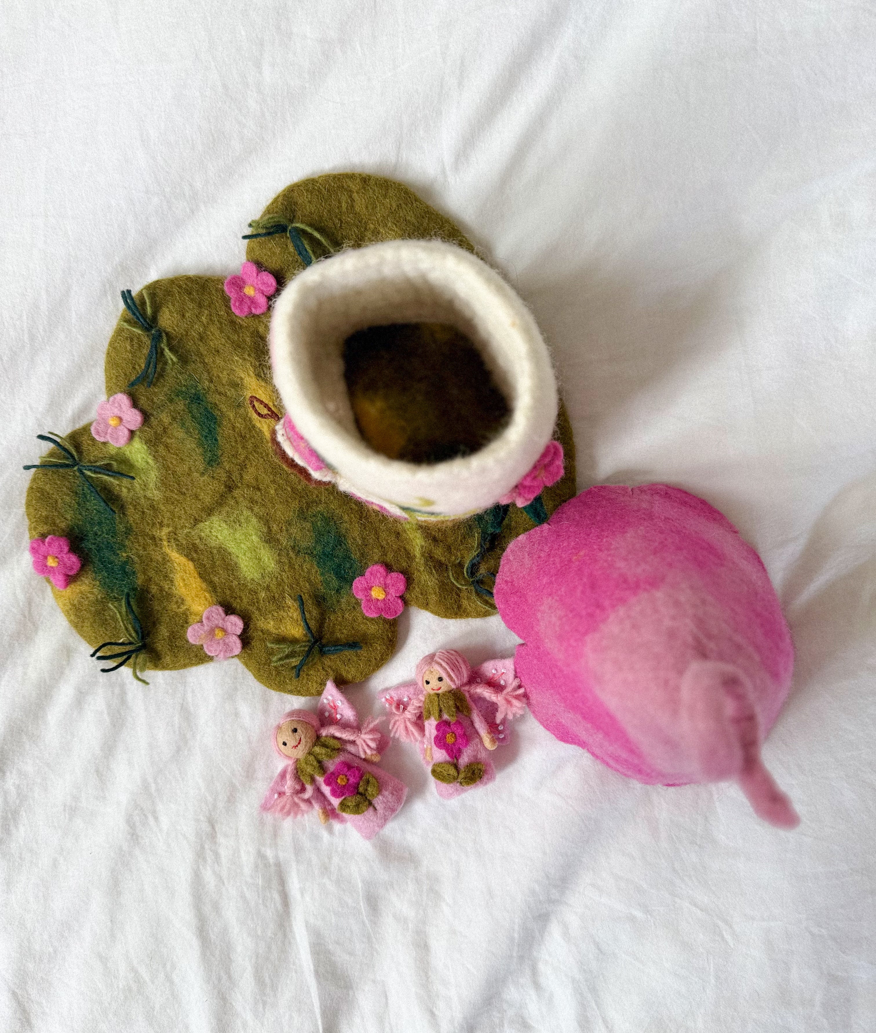 A felted wool baby bootie in the shape of a flower, shown with its matching pink felted wool hood and small pink floral accessories on a white fabric background.