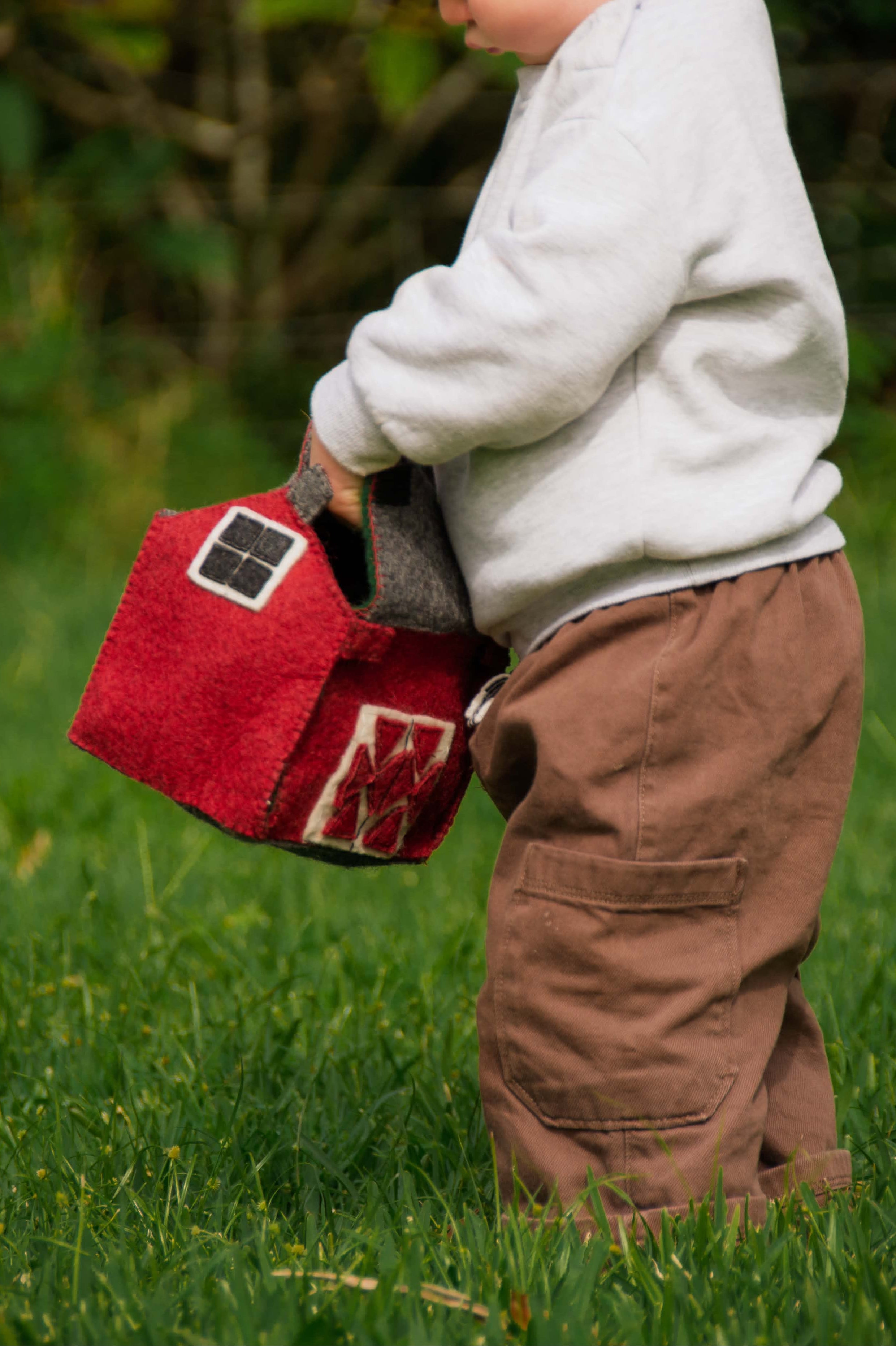 Child holding a red toy barn in a grassy outdoor setting