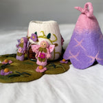 A handmade felt craft set featuring a small fairy figurine, a white felt cup decorated with colorful floral embroidery, a purple and pink gradient felt tent, and a felted green ground mat with embroidered flowers, all arranged on a white fabric background.