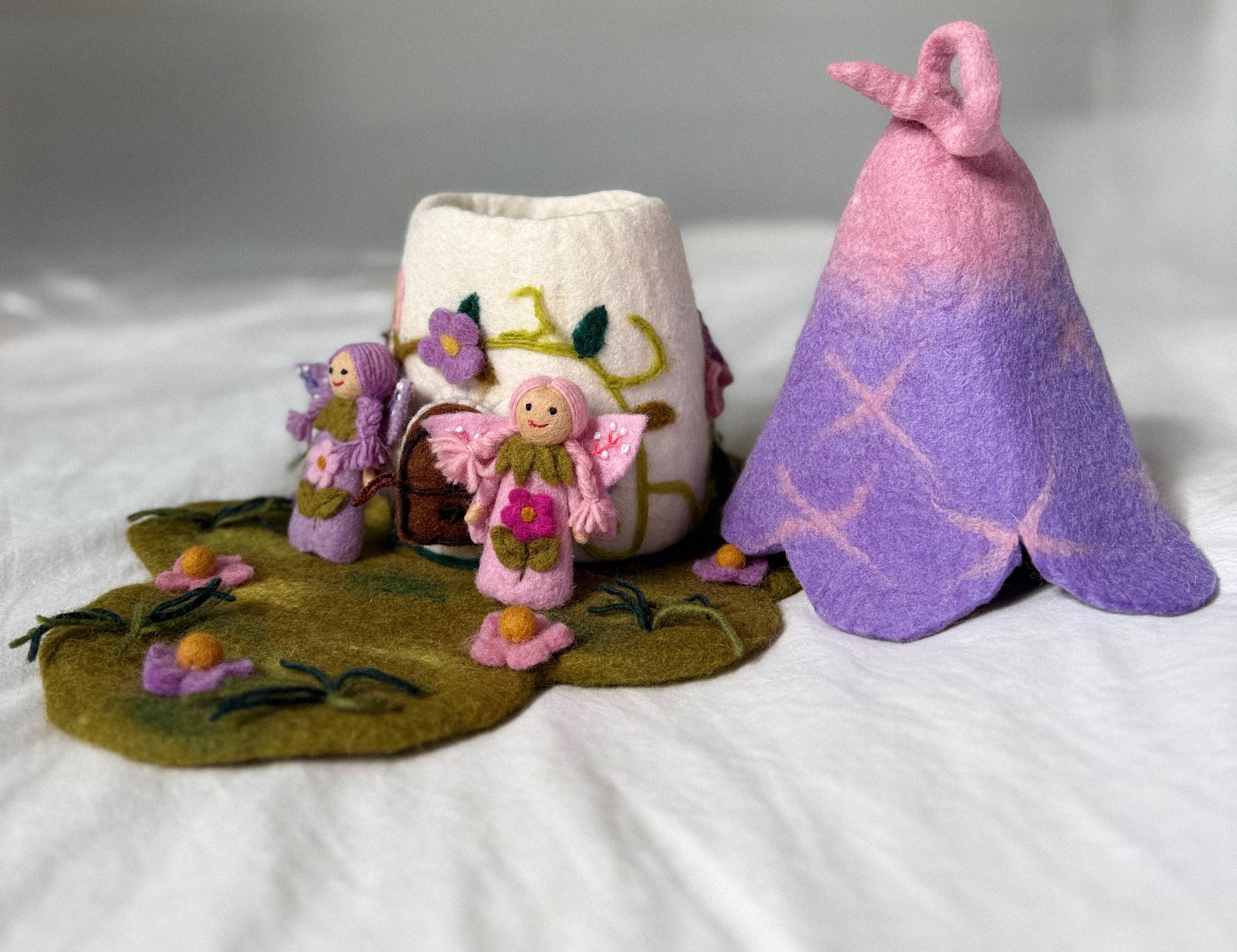 A handmade felt craft set featuring a small fairy figurine, a white felt cup decorated with colorful floral embroidery, a purple and pink gradient felt tent, and a felted green ground mat with embroidered flowers, all arranged on a white fabric background.