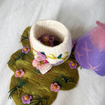 A handmade felted wool bunny cup with a white body and pink floral accents, sitting on a green felted leaf-shaped mat with pink flowers. A small pink bunny figure is tucked inside the cup, and a purple and pink felted wool object is partially visible on the right, all on a white fabric background.