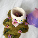 A handmade felted wool bunny cup with a white body and pink floral accents, sitting on a green felted leaf-shaped mat with pink flowers. A small pink bunny figure is tucked inside the cup, and a purple and pink felted wool object is partially visible on the right, all on a white fabric background.