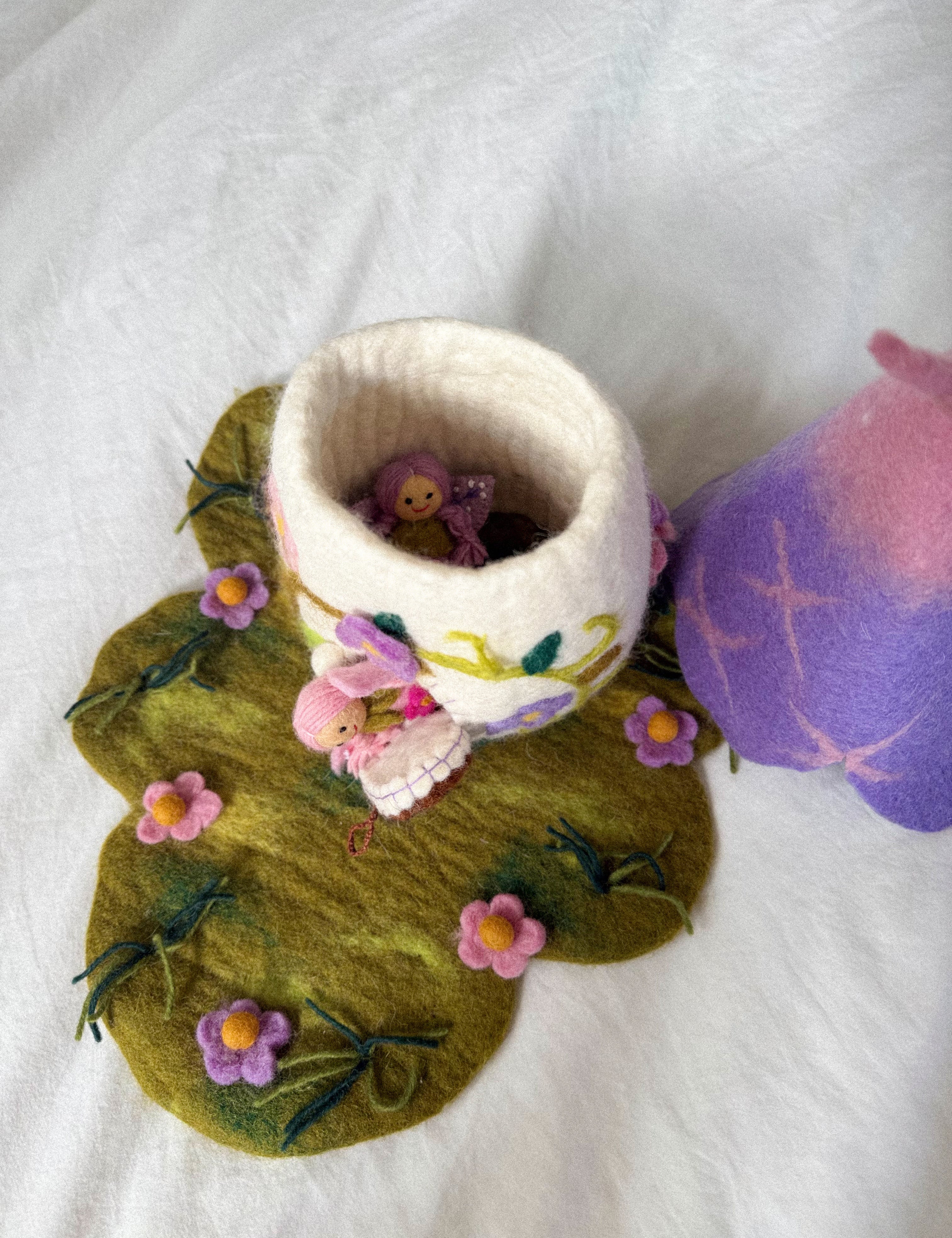 A handmade felted wool bunny cup with a white body and pink floral accents, sitting on a green felted leaf-shaped mat with pink flowers. A small pink bunny figure is tucked inside the cup, and a purple and pink felted wool object is partially visible on the right, all on a white fabric background.