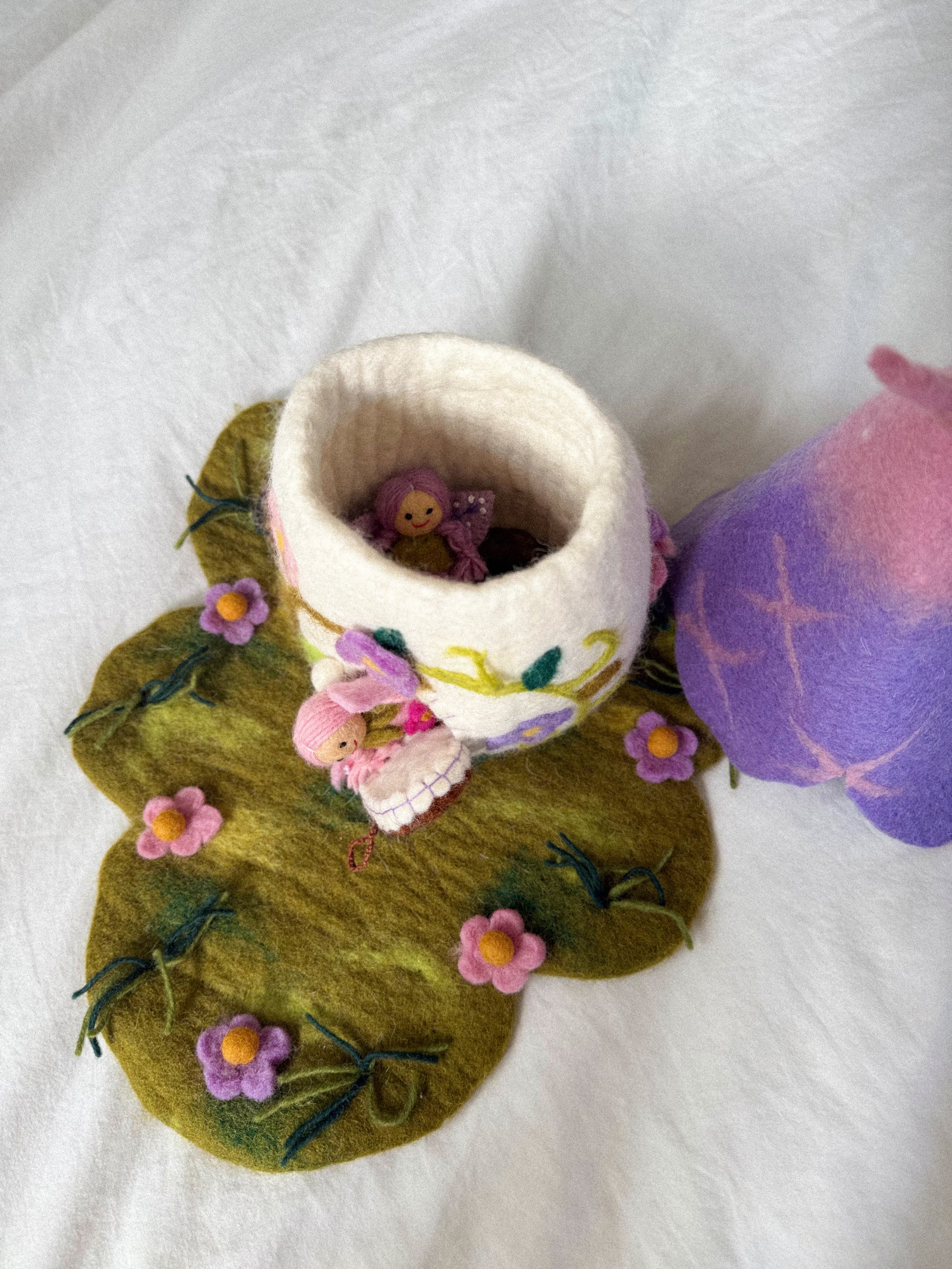 A handmade felted wool bunny cup with a white body and pink floral accents, sitting on a green felted leaf-shaped mat with pink flowers. A small pink bunny figure is tucked inside the cup, and a purple and pink felted wool object is partially visible on the right, all on a white fabric background.