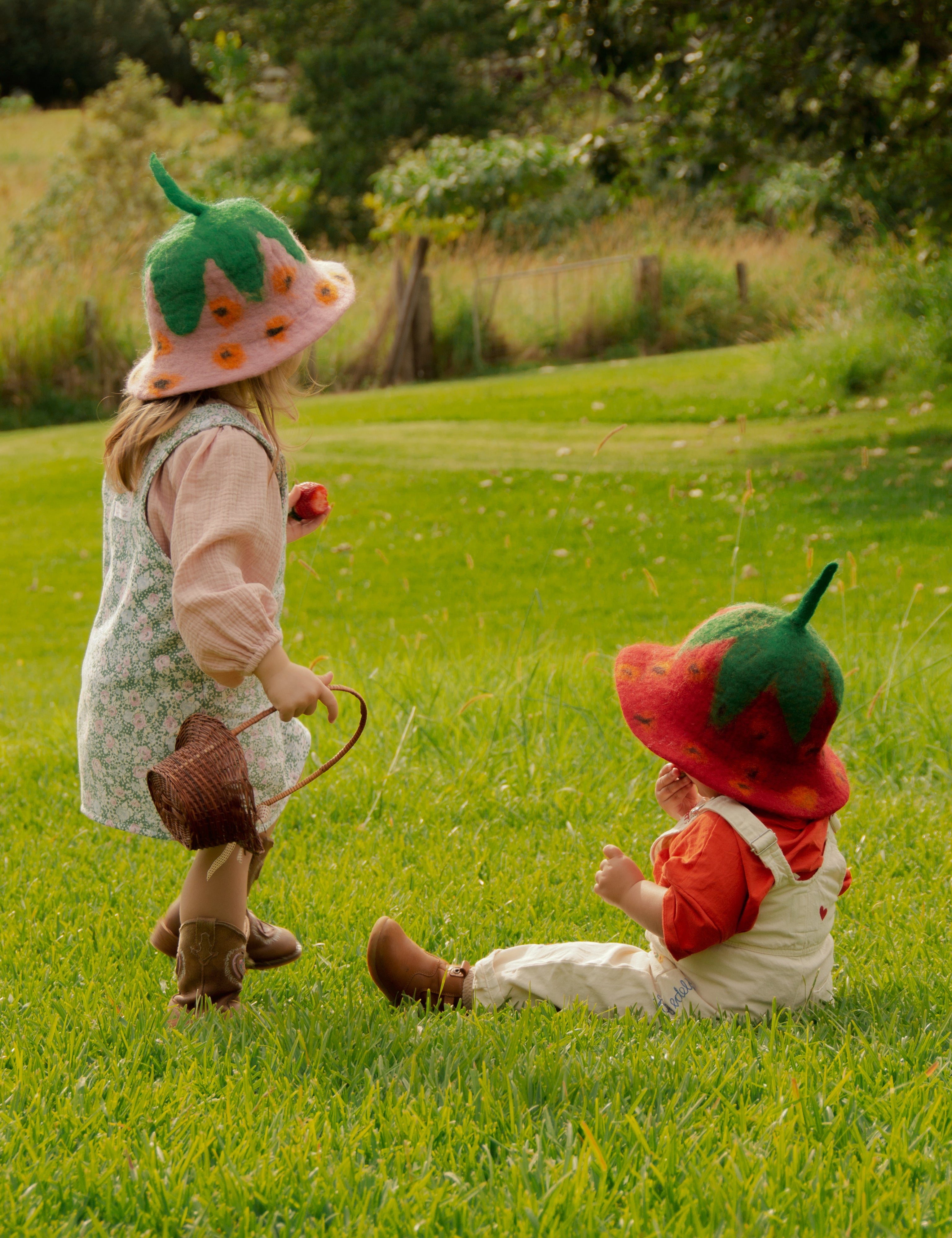 Two children in strawberry hats playing in a grassy field.
