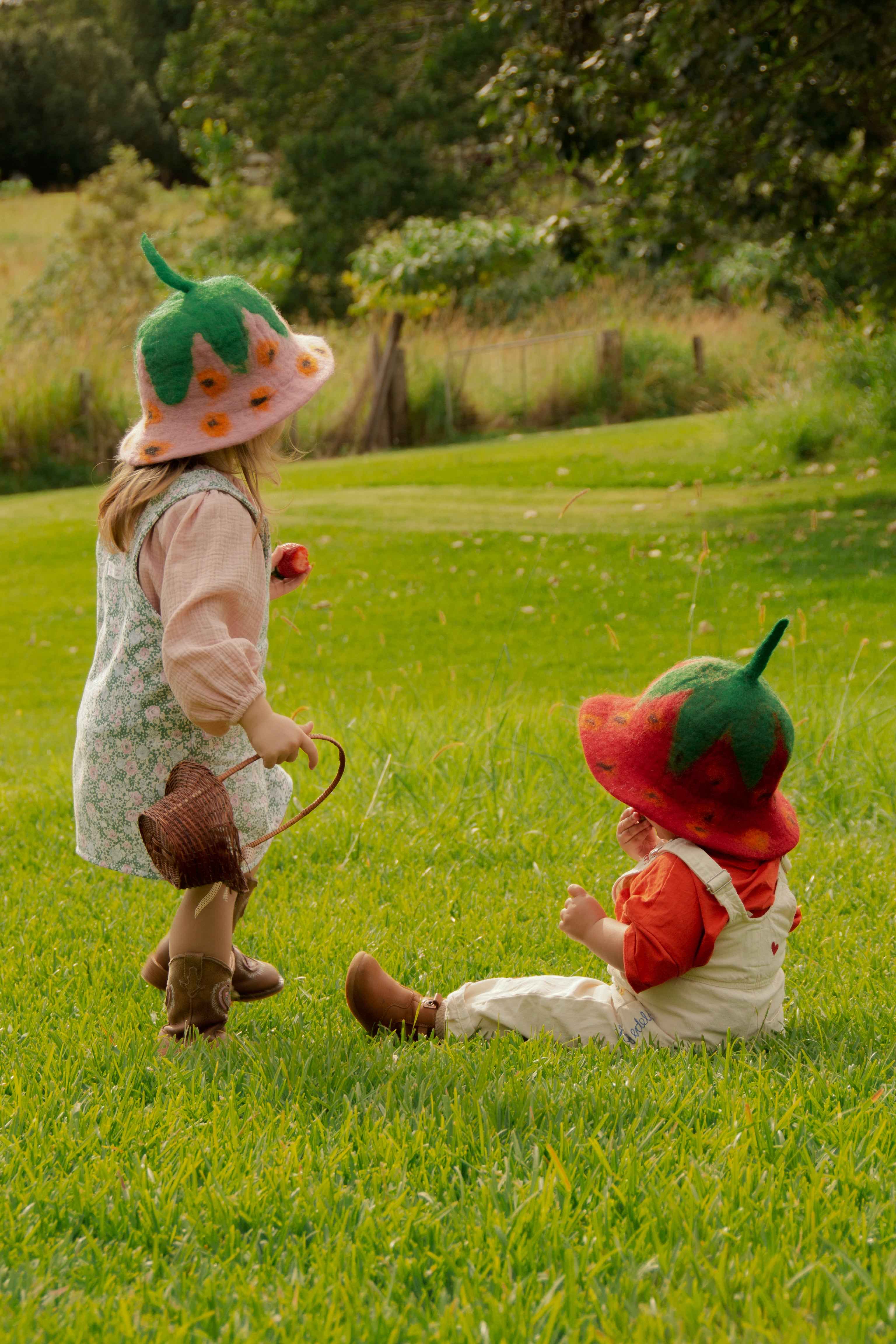 Two children in strawberry hats playing in a grassy field.