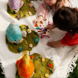 Two children playing with felted garden creatures on a white blanket outdoors.