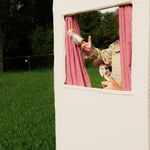 Person holding a dog and a bird in front of a small window with red checkered curtains, outdoors.