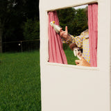 Person holding a dog and a bird in front of a small window with red checkered curtains, outdoors.