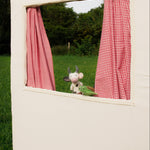 Small white dog peeking out from a window with red checkered curtains in a grassy outdoor setting.