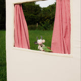 Small white dog peeking out from a window with red checkered curtains in a grassy outdoor setting.