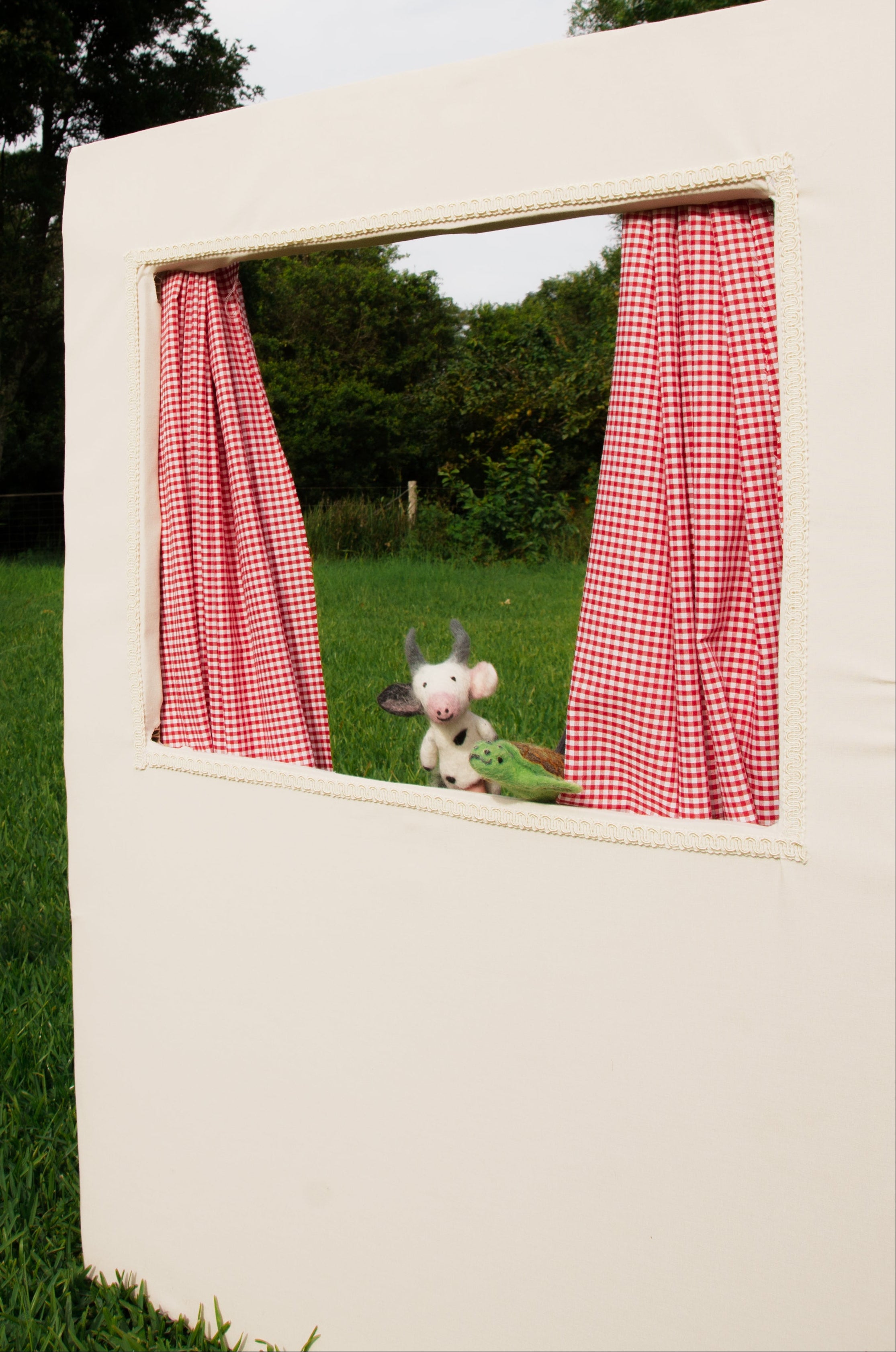 Small white dog peeking out from a window with red checkered curtains in a grassy outdoor setting.